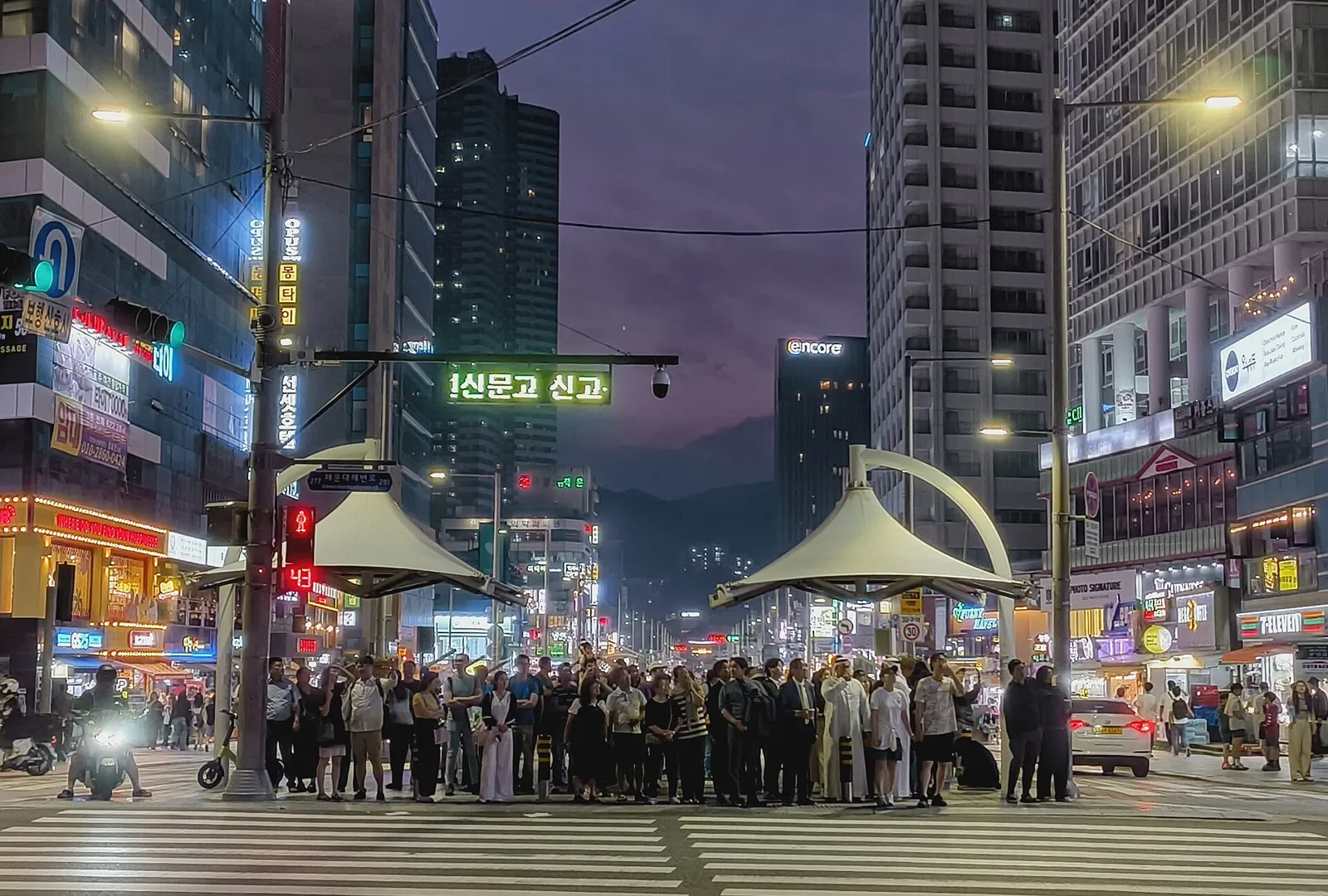 People waiting at a crosswalk on Gunam-ro in Haeundae at dusk, with lit signs, high-rises and the street's white canopies