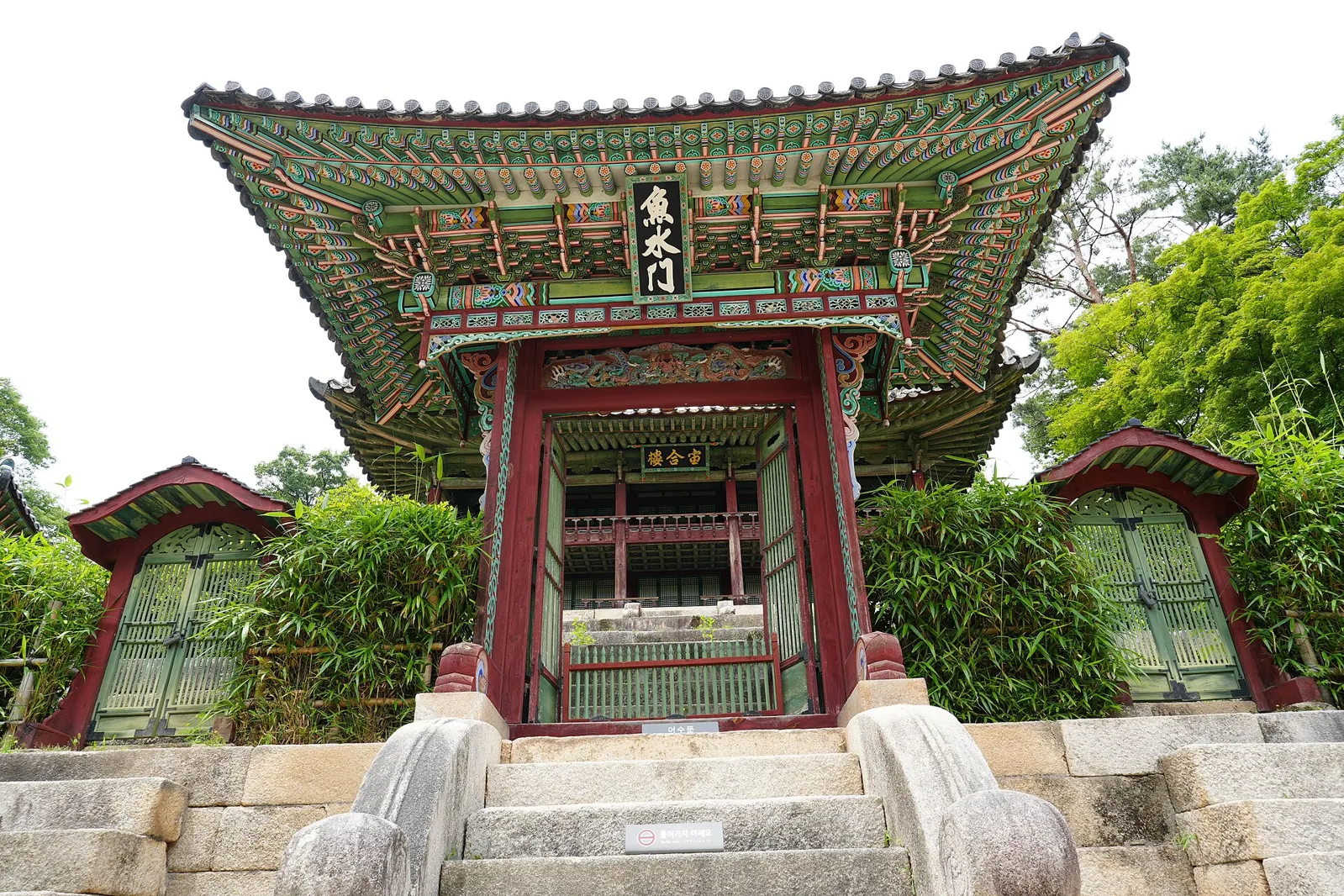 A small red gate with a painted, curved tile roof and green-and-red decoration, set between bamboo hedges at the top of stone steps