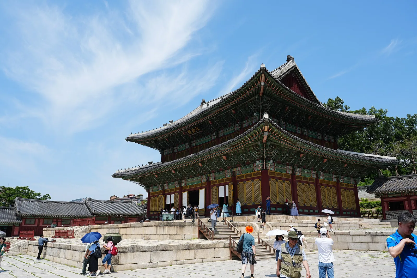A large two-storey wooden throne hall with a double tiled roof on a stone platform, with visitors in the courtyard under a blue sky