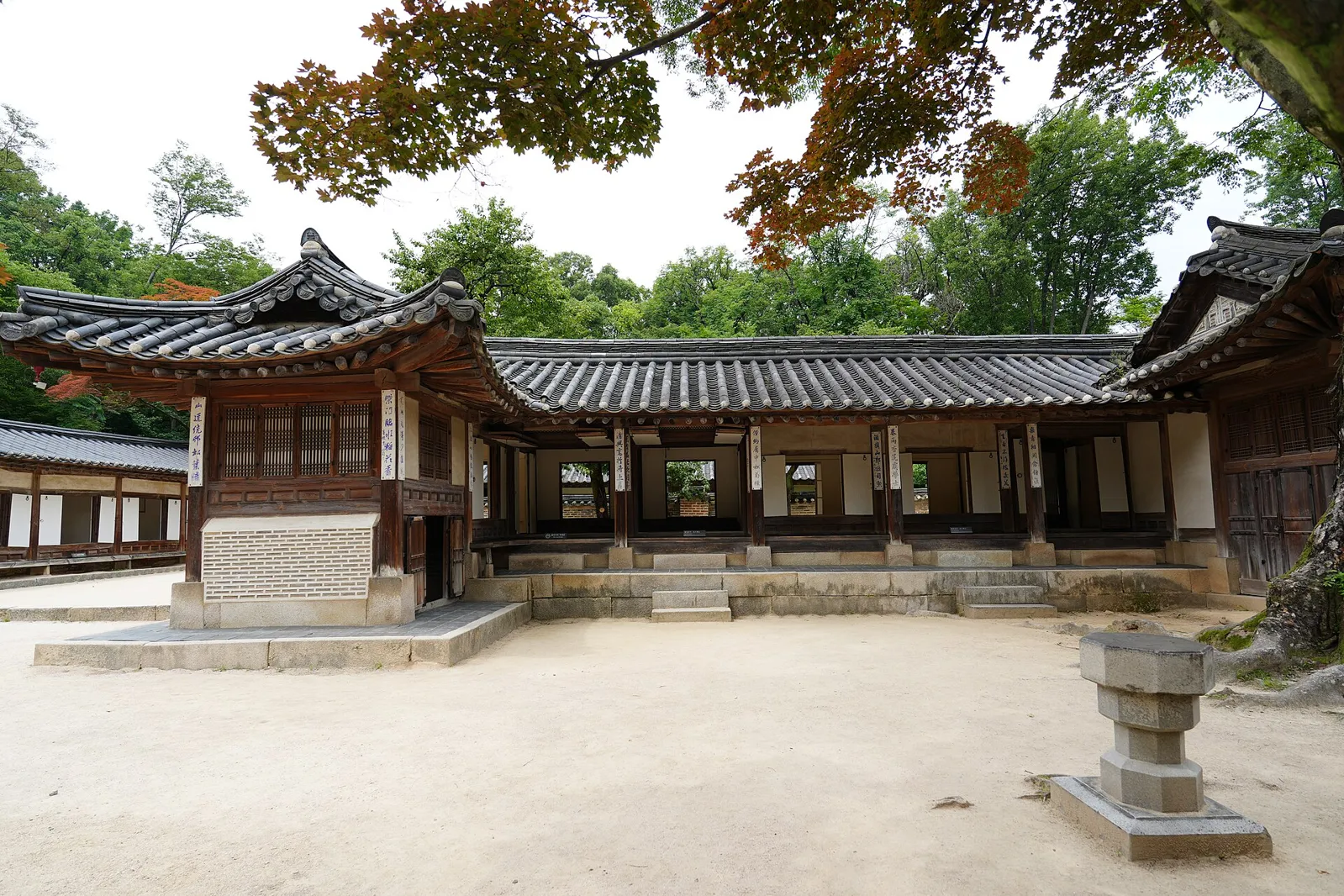 A long, unpainted wooden house with a grey tile roof around a sandy courtyard, with red maple leaves hanging in the top of the frame