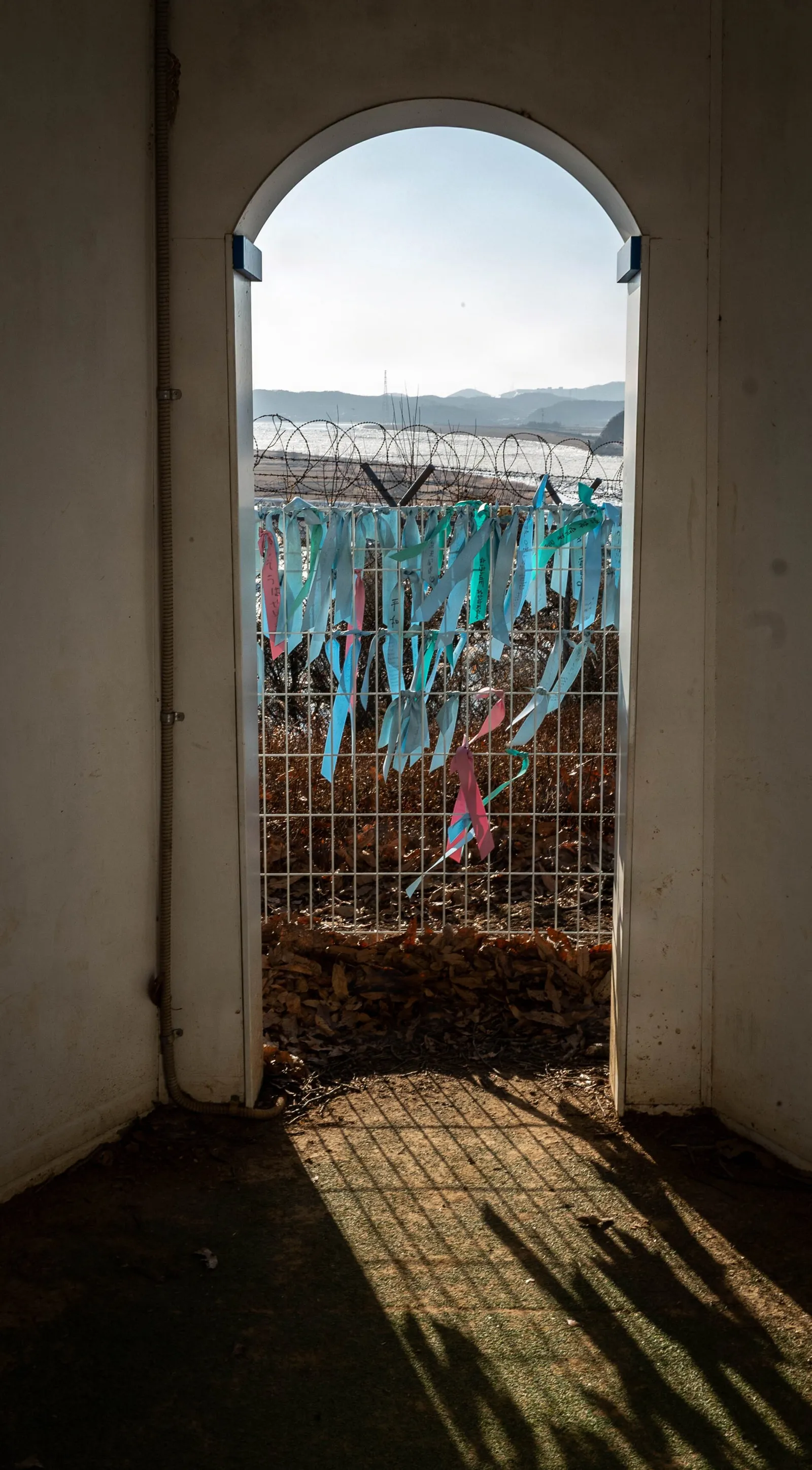 Pale blue and pink ribbons tied to a wire fence, seen through an arched doorway, with barbed wire and hills behind