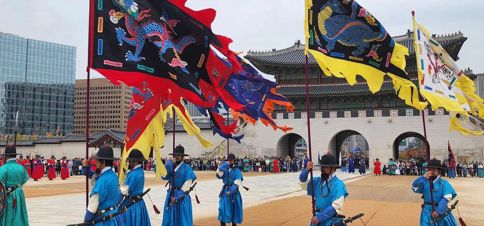 Royal guards in blue robes and black hats holding large dragon flags in front of Gwanghwamun, with a crowd watching