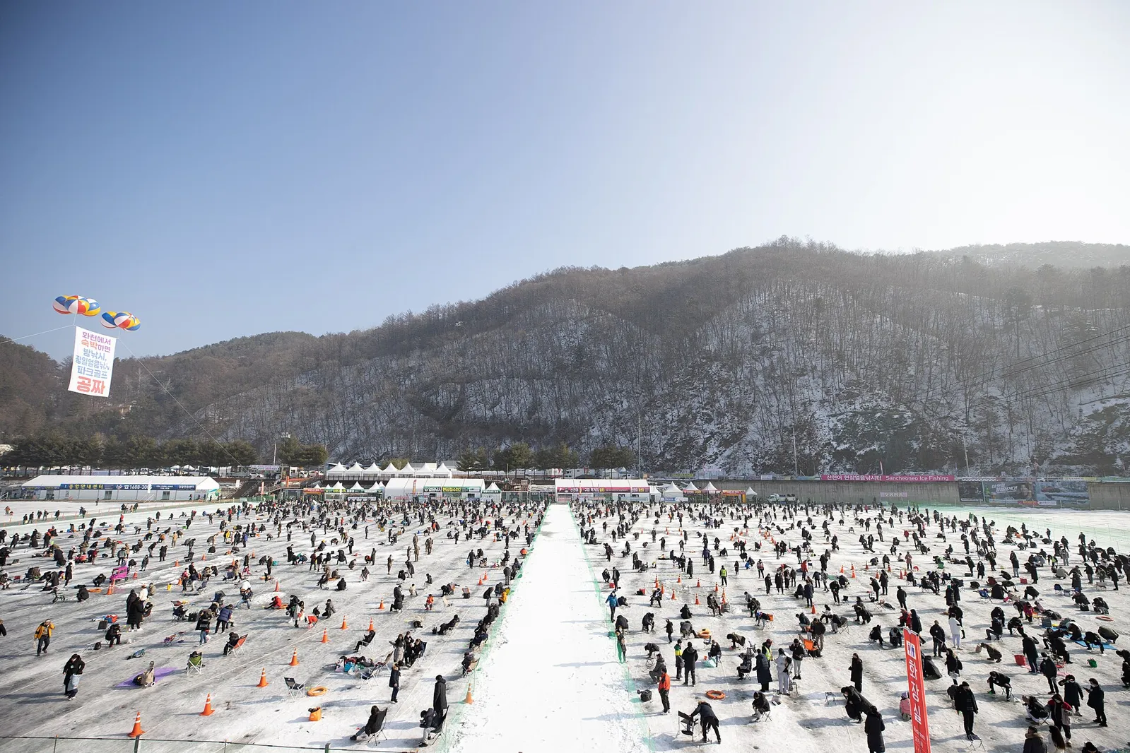 Hundreds of people ice fishing on a frozen river at the Hwacheon Sancheoneo Ice Festival, with snowy hills behind